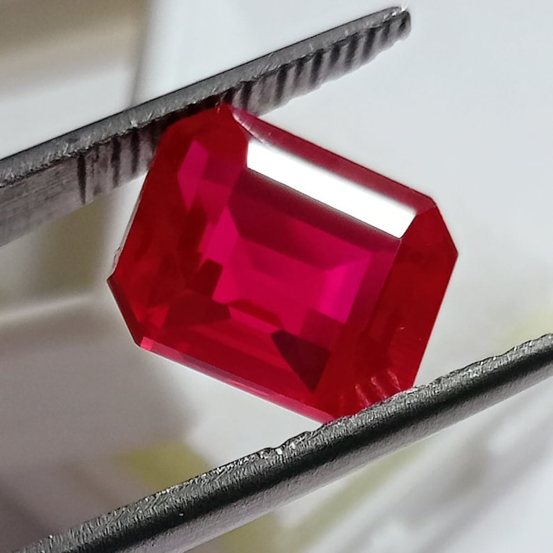 May include: A vibrant red gemstone with an emerald cut. The gemstone is held in a pair of tweezers.