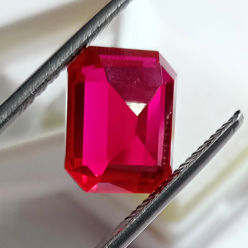 May include: A vibrant red gemstone with a square emerald cut. The gemstone is held by a pair of tweezers.