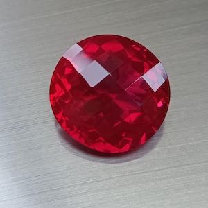 Könnte beinhalten: Ein facettierter, runder, roter Edelstein. Der Edelstein hat mehrere flache Oberflächen, die das Licht reflektieren und einen funkelnden Effekt erzeugen. Der Edelstein hat eine tiefe, satte rote Farbe.