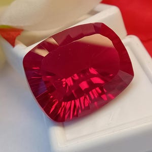 May include: A faceted, cushion-cut ruby gemstone. The gem is a deep, saturated red color with multiple facets that reflect light. The ruby is set against a white background.