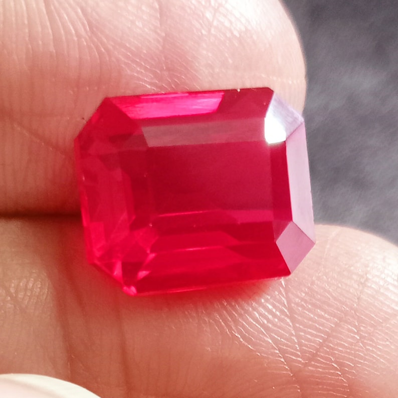 May include: A vibrant red gemstone with a rectangular emerald cut. The gemstone is faceted and has a high polish.