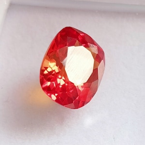 Peut inclure: Une pierre précieuse rouge de forme coussin. La pierre précieuse a une légère teinte orange et est facetée.