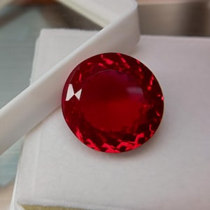 Puede incluir: Una gema redonda, facetada, de color rojo intenso. La gema está colocada sobre un fondo blanco y se sujeta en una bandeja blanca. Las facetas de la gema reflejan la luz, creando un brillo brillante. La gema es una pieza vibrante y llamativa.