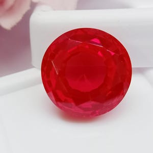 May include: A round, faceted, bright red gemstone. The gem is set against a white background, with a hint of pink in the upper left corner. The gem's surface reflects light, showcasing its many facets.