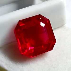 May include: A deep red gemstone with a square emerald cut. The gemstone is on a white surface.