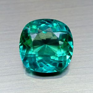 Puede incluir: Una gema tallada en cojín de color turquesa vibrante. Las facetas de la gema reflejan la luz, creando un efecto brillante. La gema se encuentra sobre un fondo metálico neutro.