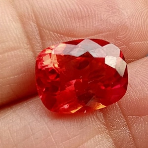 May include: A vibrant red gemstone with a cushion cut and a faceted finish. The gemstone is set in a clear, transparent setting.