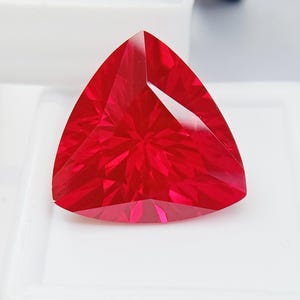 May include: A faceted, triangular-shaped gemstone with a vibrant red hue. The gem's surface reflects light, showcasing intricate internal patterns. The gemstone is set against a white background, highlighting its color and shape. This is a gemstone.