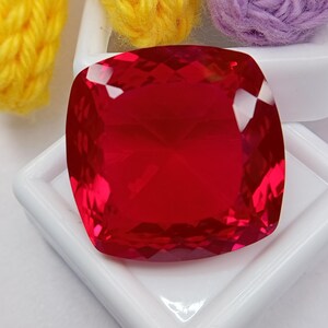 Puede incluir: Una gema roja vibrante de corte cojín con una superficie facetada que refleja la luz. La gema se muestra en un contenedor cuadrado blanco, colocada sobre un fondo con elementos de hilo texturizado en colores amarillo y morado.