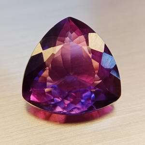 May include: A faceted, triangular gemstone with a gradient of colors, transitioning from deep purple to pink and blue. The gem is set against a neutral background, showcasing its intricate cuts and reflective surfaces.