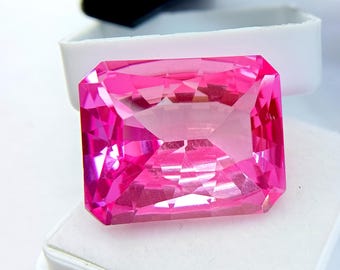 May include: A faceted, rectangular-cut, bright pink gemstone. The gem is set against a white background, with a white container visible in the background. The gem's facets reflect light, creating a sparkling effect. The gem is a vibrant shade of pink.