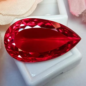 May include: A pear-shaped, faceted gemstone with a vibrant, deep red color. The stone features a brilliant cut with numerous triangular and kite-shaped facets that reflect light across its surface, resting on a small, square white display base.