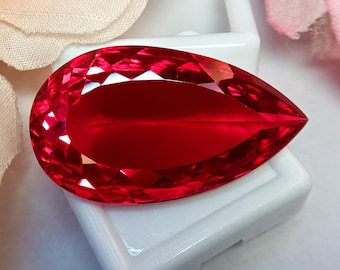 May include: A pear-shaped, faceted gemstone with a vibrant, deep red color. The stone features a brilliant cut with numerous triangular and kite-shaped facets that reflect light across its surface, resting on a small, square white display base.