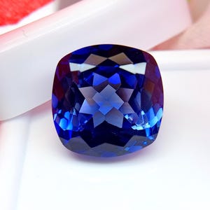 Puede incluir: Una gema facetada, de talla cojín, de color azul intenso. La superficie de la gema refleja la luz, mostrando un complejo patrón de facetas. La gema se encuentra sobre un fondo blanco, resaltando su color y claridad.
