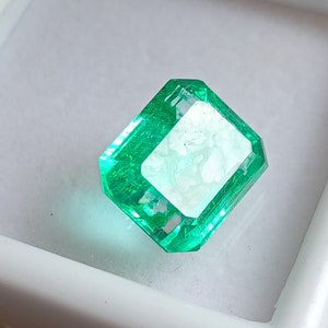 Puede incluir: Una gema de esmeralda verde vibrante con un corte facetado. La esmeralda está engastada en un engaste blanco transparente.