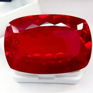 Pode incluir: Uma grande pedra preciosa de rubi facetada com corte almofada. A gema é de cor vermelha profunda e saturada com alto grau de clareza. O rubi está colocado sobre uma superfície branca, e as facetas refletem a luz.