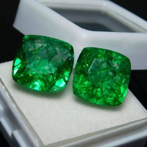 May include: Two emerald green gemstones, cut into a square shape with rounded corners, are displayed in a white box. The gemstones are translucent and have a slight sparkle.