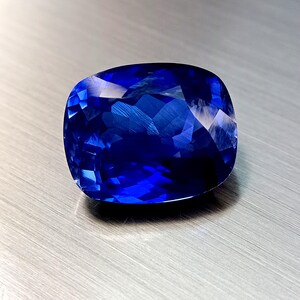 Peut inclure: Une grosse pierre pr&eacute;cieuse saphir de taille coussin, d'un bleu profond et vibrant. Les facettes refl&egrave;tent la lumi&egrave;re, cr&eacute;ant un effet scintillant. La pierre est pos&eacute;e sur une surface m&eacute;tallique bross&eacute;e.