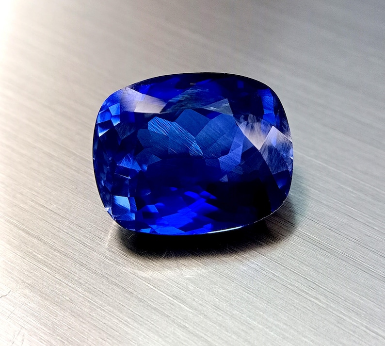 Peut inclure: Une grosse pierre pr&eacute;cieuse saphir de taille coussin, d'un bleu profond et vibrant. Les facettes refl&egrave;tent la lumi&egrave;re, cr&eacute;ant un effet scintillant. La pierre est pos&eacute;e sur une surface m&eacute;tallique bross&eacute;e.