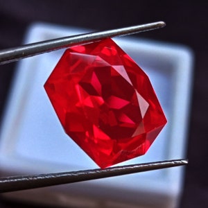 May include: A deep red gemstone with a faceted cut, held by tweezers against a white background.
