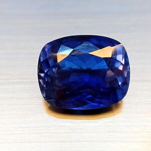 Peut inclure: Une pierre pr&eacute;cieuse taill&eacute;e en coussin de couleur bleu fonc&eacute;. La surface de la gemme refl&egrave;te la lumi&egrave;re, r&eacute;v&eacute;lant sa structure interne et ses facettes. La pierre est pos&eacute;e sur une surface claire, mettant en valeur sa couleur et sa clart&eacute;.