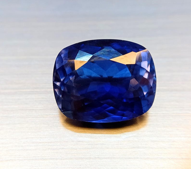 Peut inclure: Une pierre pr&eacute;cieuse taill&eacute;e en coussin de couleur bleu fonc&eacute;. La surface de la gemme refl&egrave;te la lumi&egrave;re, r&eacute;v&eacute;lant sa structure interne et ses facettes. La pierre est pos&eacute;e sur une surface claire, mettant en valeur sa couleur et sa clart&eacute;.
