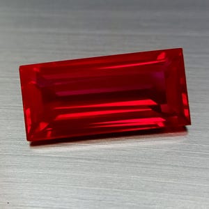 Puede incluir: Una gema rectangular y facetada de color rojo intenso. La gema tiene una superficie pulida con reflejos internos, creando una apariencia vibrante. La forma es un corte baguette clásico, que muestra la claridad y el color de la gema.