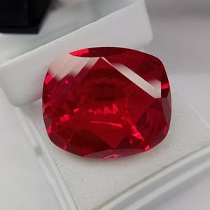 May include: A faceted, cushion-cut, bright red gemstone. The gem is set in a white display box, with a black object visible in the background. The gemstone's facets reflect light, creating a brilliant appearance. The overall theme is jewelry and gemstones.
