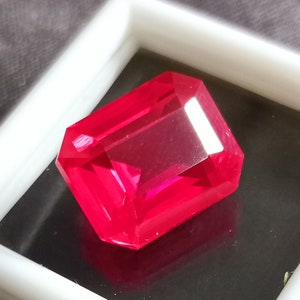 May include: A vibrant red gemstone with a rectangular emerald cut. The gemstone is displayed in a white box.