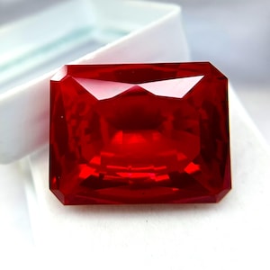 Może przedstawiać: Żywy, prostokątny, czerwony kamień szlachetny. Kamień jest fasetowany, odbija światło i ukazuje swój głęboki kolor. Prezentowany jest na białej powierzchni, być może w pudełku na biżuterię, podkreślając jego blask. Kształt kamienia szlachetnego to idealny kwadrat.
