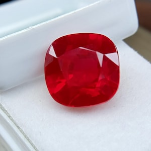 May include: A cushion-cut, vibrant red gemstone, likely a ruby, displayed in a white box. The gem's facets reflect light, showcasing its clarity and rich color. The square shape with rounded corners is prominent.
