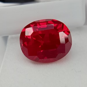 Peut inclure: Une pierre précieuse rouge foncé, taillée en forme ovale et à facettes. La gemme possède de multiples facettes qui réfléchissent la lumière, créant un éclat brillant. La pierre est posée sur un fond blanc, soulignant sa couleur riche et sa clarté. Cette pierre est adaptée à la joaillerie.
