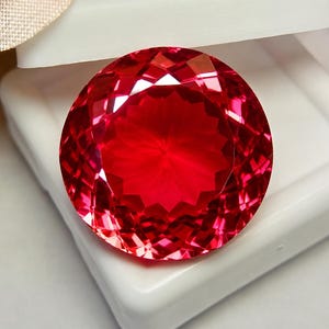 May include: A round, faceted gemstone with a vibrant, deep red color. The stone features a complex, symmetrical cut with numerous triangular and trapezoidal facets that create a brilliant, reflective surface. It is displayed inside a white box.