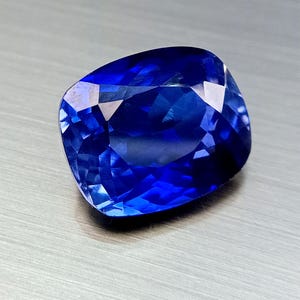 Peut inclure: Une pierre pr&eacute;cieuse bleu profond taill&eacute;e en coussin, avec des facettes r&eacute;fl&eacute;chissant la lumi&egrave;re. Le saphir est d'une couleur vive et satur&eacute;e, et la taille met en valeur son &eacute;clat. La gemme est pos&eacute;e sur un fond neutre et textur&eacute;.