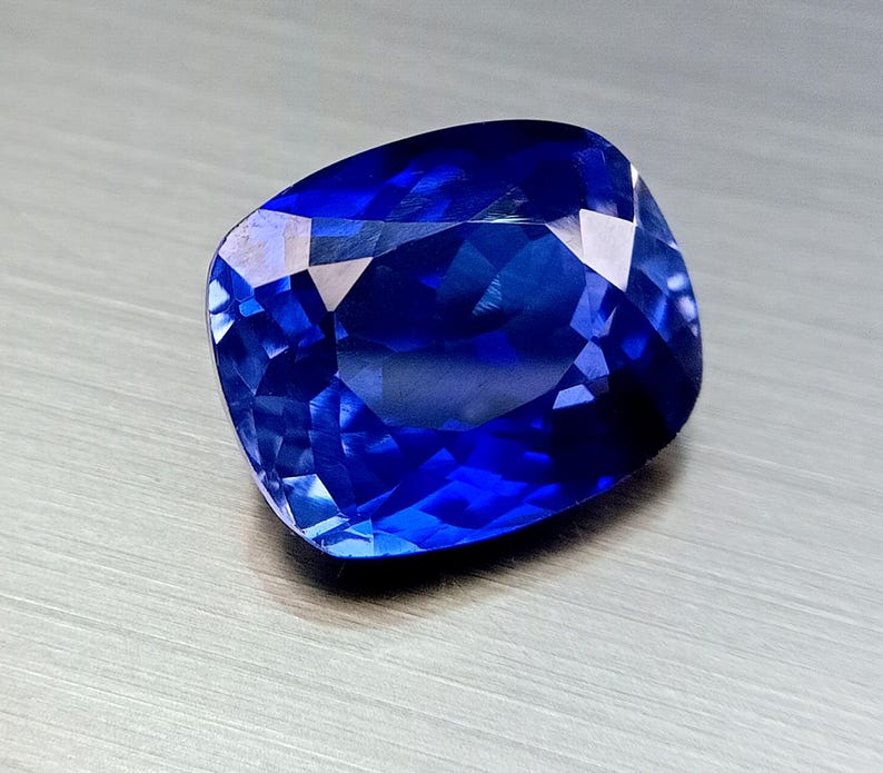 Peut inclure: Une pierre pr&eacute;cieuse bleu profond taill&eacute;e en coussin, avec des facettes r&eacute;fl&eacute;chissant la lumi&egrave;re. Le saphir est d'une couleur vive et satur&eacute;e, et la taille met en valeur son &eacute;clat. La gemme est pos&eacute;e sur un fond neutre et textur&eacute;.