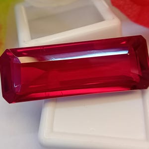 Puede incluir: Una gema de rubí rectangular y facetada, de color rojo intenso. La gema se muestra sobre un fondo blanco, resaltando su claridad y brillo. El corte de la piedra destaca sus facetas, reflejando la luz. Una hermosa pieza para joyería.