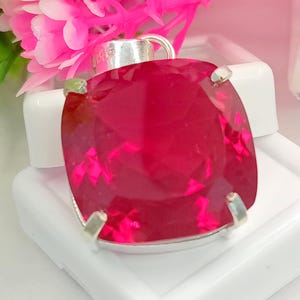 May include: A square-cut, faceted deep pink gemstone pendant set in a silver-toned metal prong setting. The pendant features a smooth, reflective surface with sharp edges and a metallic bail at the top for attachment to a chain or cord.