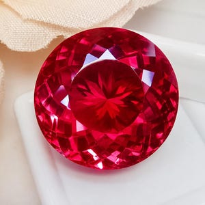 May include: A round, faceted gemstone with a vibrant, deep red color. The stone features a complex, geometric cut that creates numerous triangular and diamond-shaped facets, reflecting light to produce a brilliant, sparkling appearance throughout.