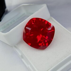 May include: A faceted, cushion-cut, deep red gemstone rests in a white jewelry box. The gem's surface reflects light, showcasing its internal structure. The box is open, revealing the gem's vibrant color and clarity.