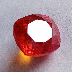 May include: A red gemstone with a faceted cut. The gemstone is a cushion shape and has a high polish.