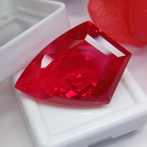 Puede incluir: Una gema de color rojo intenso y vibrante con un corte facetado en forma de escudo. La piedra muestra ángulos geométricos definidos y una superficie brillante y reflectante, colocada dentro de una pequeña caja de presentación cuadrada blanca.
