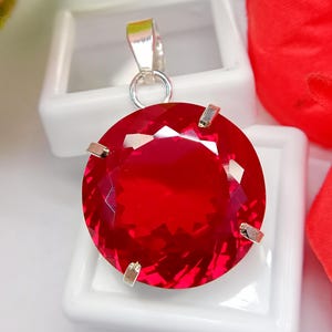 May include: A round, faceted, vibrant red gemstone pendant set in a silver-toned metal prong setting. The circular stone features a brilliant cut with multiple reflective surfaces, attached to a simple silver-toned metal bail at the top.