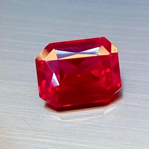 Puede incluir: Una gema roja vibrante, tallada en forma rectangular. La gema tiene un alto grado de claridad, reflejando la luz y mostrando su estructura interna. Los bordes están cortados con precisión, creando una forma simétrica. La gema se encuentra sobre un fondo neutro.