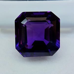 May include: A faceted, rectangular-cut amethyst gemstone. The gem is a deep, saturated purple color with sharp, clean facets that reflect light. The gemstone is set against a neutral background, highlighting its color and clarity.