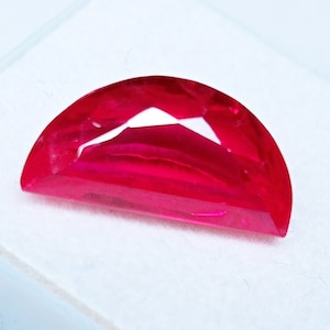May include: A bright red, half-moon shaped gemstone with multiple facets. The gem has a flat base and a curved top, creating a play of light and shadow. The gemstone is set against a white background. The gem is likely a ruby.