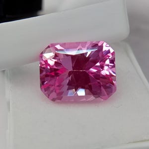 May include: A vibrant, rectangular-cut pink gemstone with multiple facets. The gem's colour is a bright pink, with facets that reflect light. The gemstone is set against a white background, highlighting its colour and clarity. A beautiful addition to any jewellery collection.