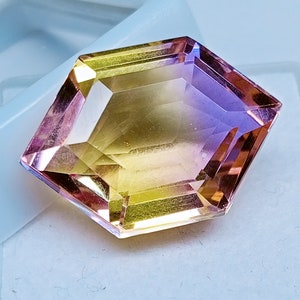 May include: A faceted gemstone with a yellow to purple gradient. The gemstone is cut in a hexagonal shape with beveled edges.
