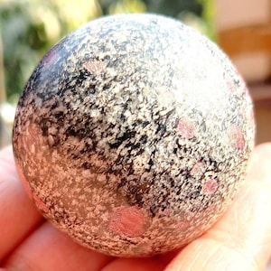 May include: A polished, spherical stone with a speckled pattern of black, white, and reddish-pink spots. The stone has a smooth, rounded shape and a slightly reflective surface, suggesting it is a decorative item or a collectible.
