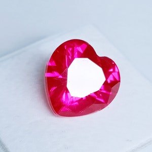 May include: A heart-shaped, faceted gemstone in a vibrant shade of red. The gemstone has a bright, reflective surface and is set against a white background. The cut of the stone creates multiple facets.