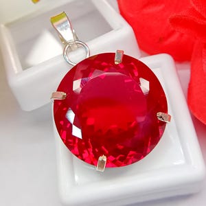 Puede incluir: Un colgante de piedra preciosa roja vibrante, redondo y facetado, montado en un engaste de metal plateado con garras. El colgante presenta un corte circular con múltiples facetas que reflejan la luz, unido a una anilla de metal plateado en la parte superior.